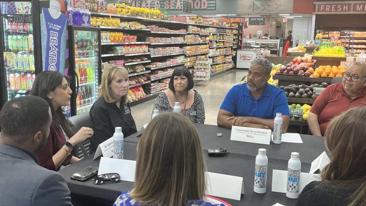 Congressman Steven Horsford sits at a roundtable discussion inside a grocery store, surrounded by community members and professionals. The group is engaged in conversation at a table covered with bottled drinks and name cards. Behind them, grocery shelves are stocked with colorful beverages, snacks, and produce, highlighting the store setting.
