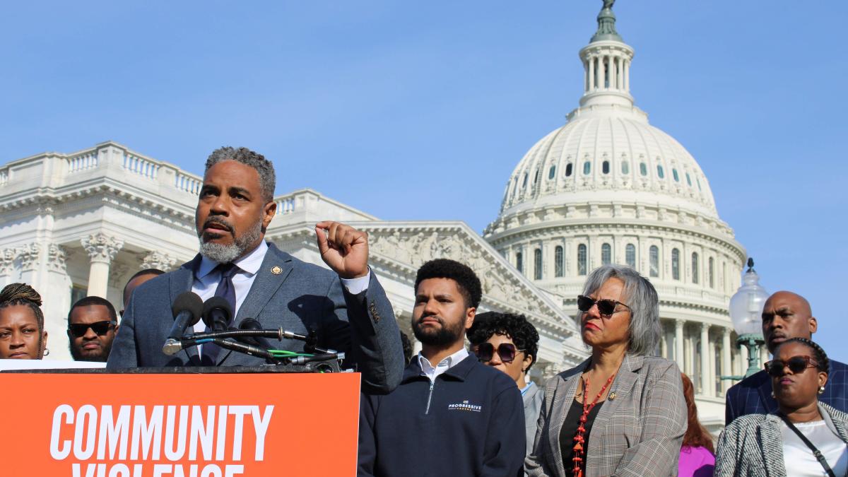 Congressman Steven Horsford speaks at a podium with 'Community Violence' written in bold white letters on an orange background. He is wearing a dark blue suit and tie, raising his fist slightly while addressing the crowd. Behind him, several individuals, including Rep. Robin Kelly and Rep. Maxwell Frost, stand in support. The U.S. Capitol building is prominently visible in the background under a clear blue sky