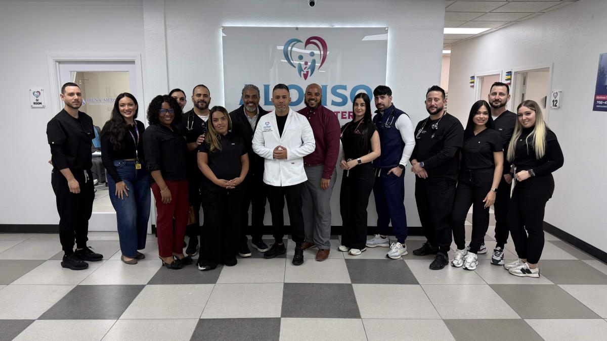A group of about 14 healthcare staff members stand together in a bright clinic hallway, posing for a photo. At the center, a man in a white medical coat stands with hands clasped, surrounded by colleagues dressed in black scrubs and business-casual attire. Behind them is a wall sign with a heart-shaped logo and the word “CareSource.” The floor has gray-and-white tiles, and fluorescent ceiling lights illuminate the space.