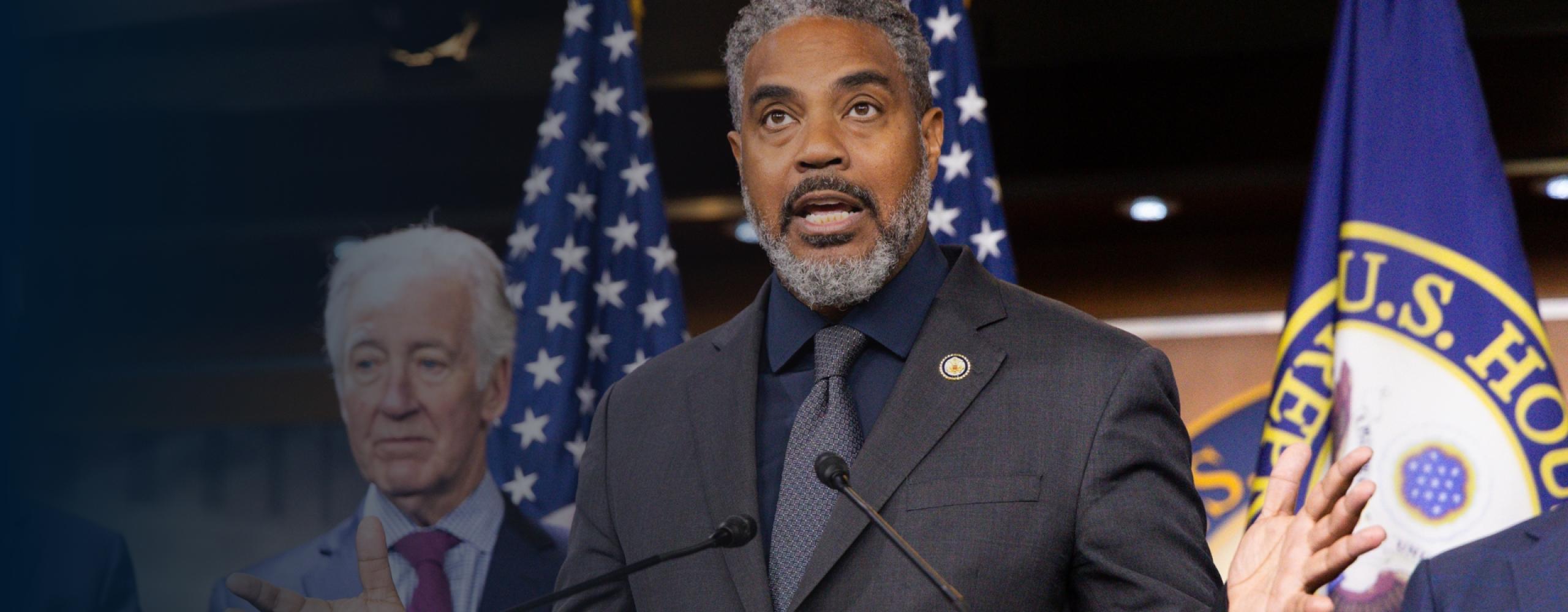 Congressman Steven Horsford speaks at a podium with microphones, gesturing with both hands. Behind him are U.S. flags and the seal of the House of Representatives. Another man stands slightly behind him, listening attentively.