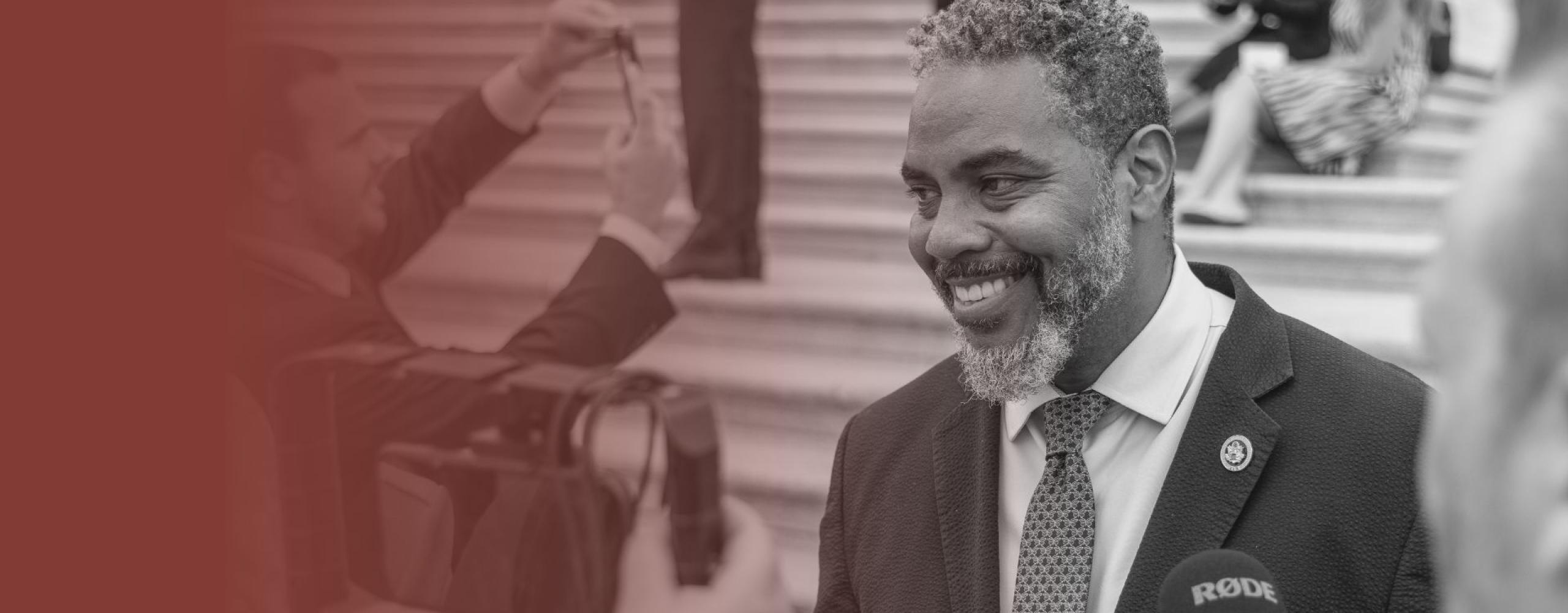 Congressman Steven Horsford smiles while speaking to reporters outside on the U.S. Capitol steps. He is wearing a dark suit and patterned tie, and is holding a conversation with several journalists, one of whom is holding a RØDE microphone. The scene is captured in black and white with a red gradient on the left side. People and media equipment are visible in the background.