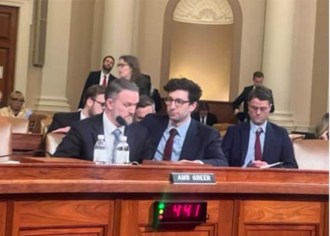 Ambassador Greer, seated on the far left, has a surprised expression on his face as he observes the proceedings. Two men in suits sit closely together at a congressional hearing table labeled "AMB GREER," with one speaking into a microphone while the other listens attentively. Two water bottles and papers are on the desk. Other attendees sit behind them in a large, formal hearing room with columns and arched architectural details. A digital timer on the desk shows "4:41.