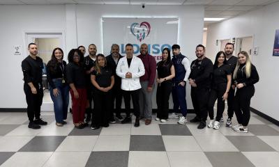 A group of about 14 healthcare staff members stand together in a bright clinic hallway, posing for a photo. At the center, a man in a white medical coat stands with hands clasped, surrounded by colleagues dressed in black scrubs and business-casual attire. Behind them is a wall sign with a heart-shaped logo and the word “CareSource.” The floor has gray-and-white tiles, and fluorescent ceiling lights illuminate the space.