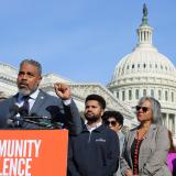 Congressman Steven Horsford speaks at a podium with 'Community Violence' written in bold white letters on an orange background. He is wearing a dark blue suit and tie, raising his fist slightly while addressing the crowd. Behind him, several individuals, including Rep. Robin Kelly and Rep. Maxwell Frost, stand in support. The U.S. Capitol building is prominently visible in the background under a clear blue sky